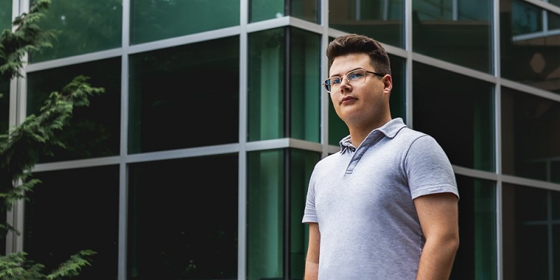 Photo of student Kyle Cook standing outside on the Saint Mary's campus. He has short brown hair, wears glasses and has a grey short-sleeved shirt on. The picture shows him from the waist up, with glass walls of a building behind him.